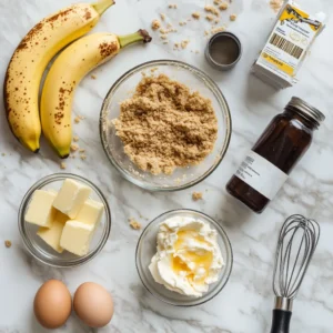 Essential ingredients for banana pudding cheesecake, including graham cracker crumbs, cream cheese, bananas, eggs, butter, sugar, vanilla extract, and banana pudding mix, arranged on a marble countertop with natural light