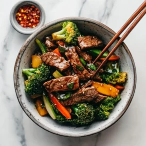 Beef stir fry served in a bowl with chopsticks and sides of soy sauce and chili flakes on a marble countertop.